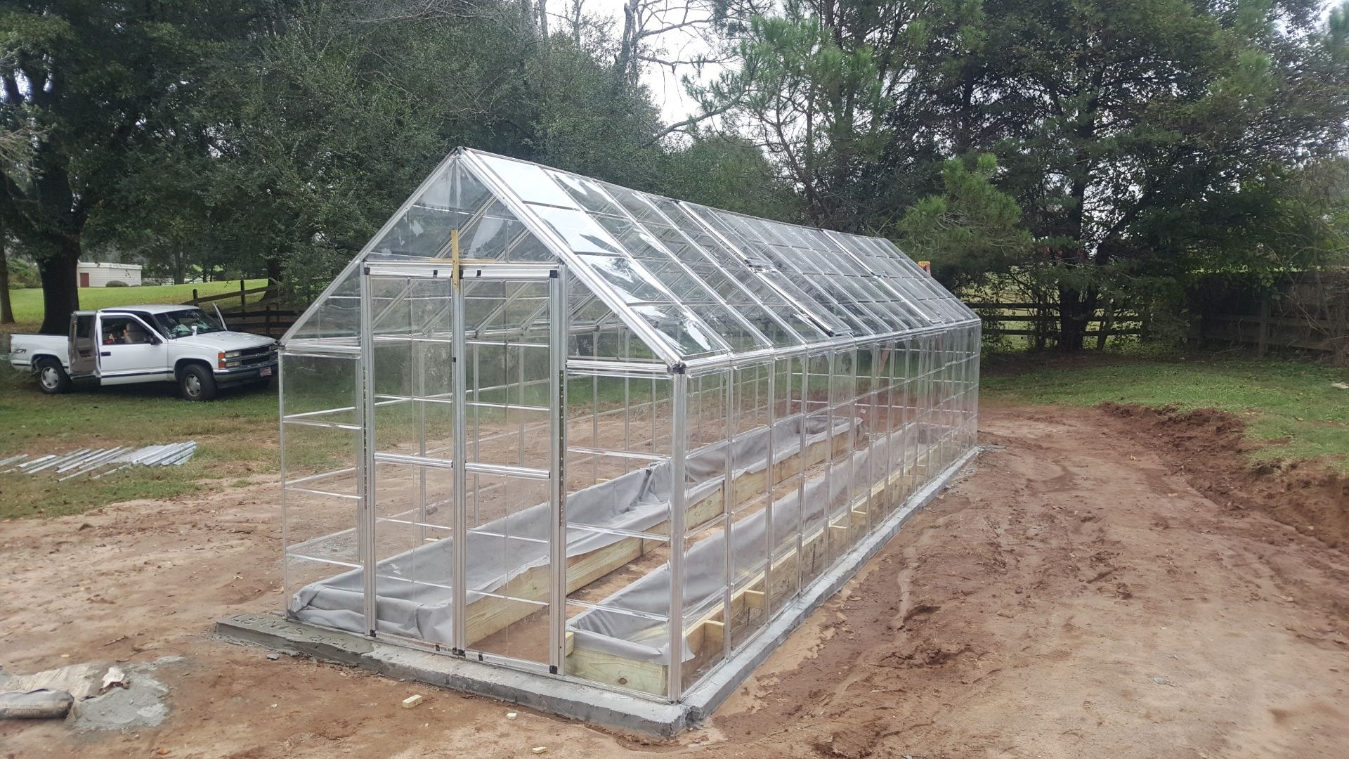 A rectangular, clear greenhouse in a muddy field with a white truck parked nearby.