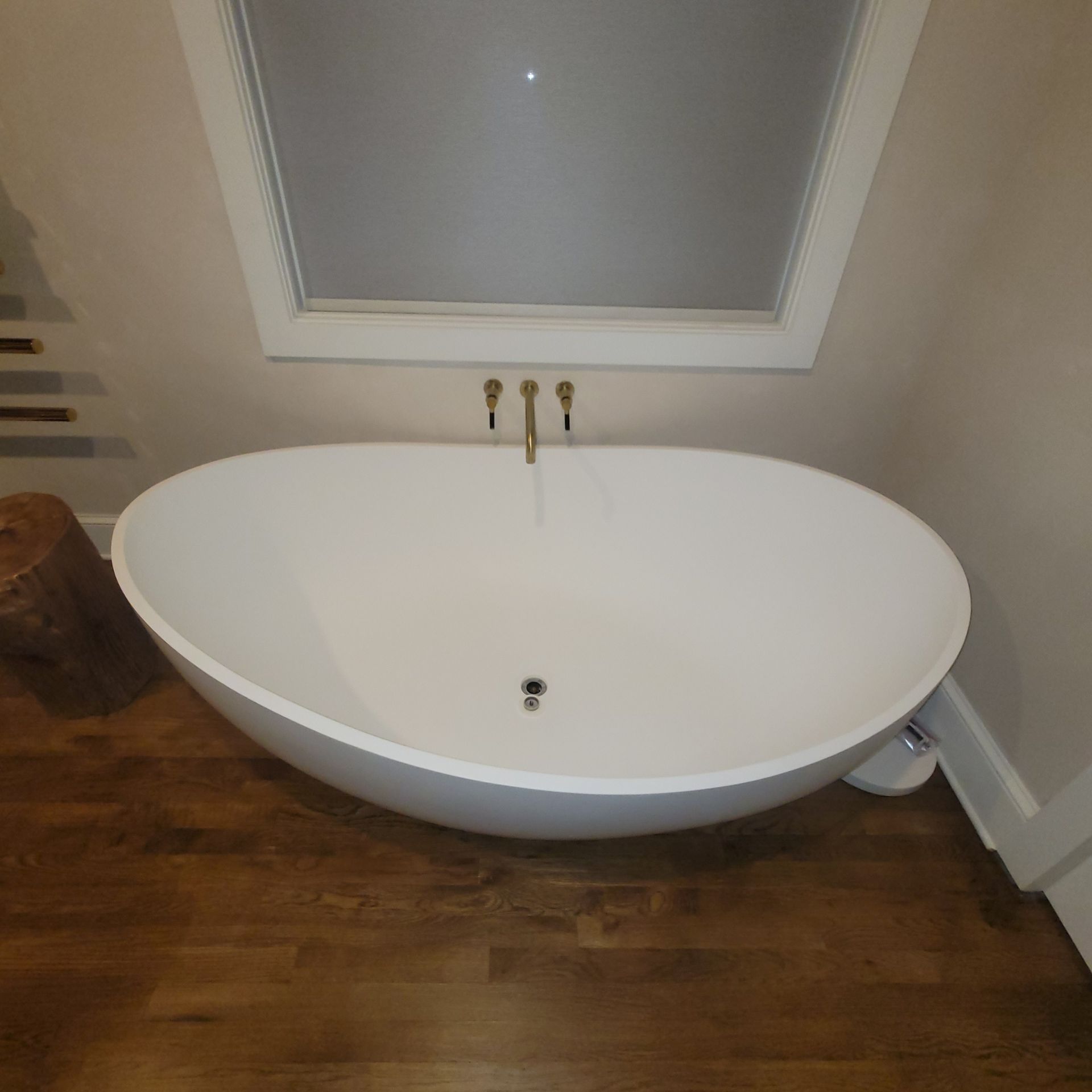 White oval bathtub under a window with gold fixtures in a bathroom with hardwood floors.