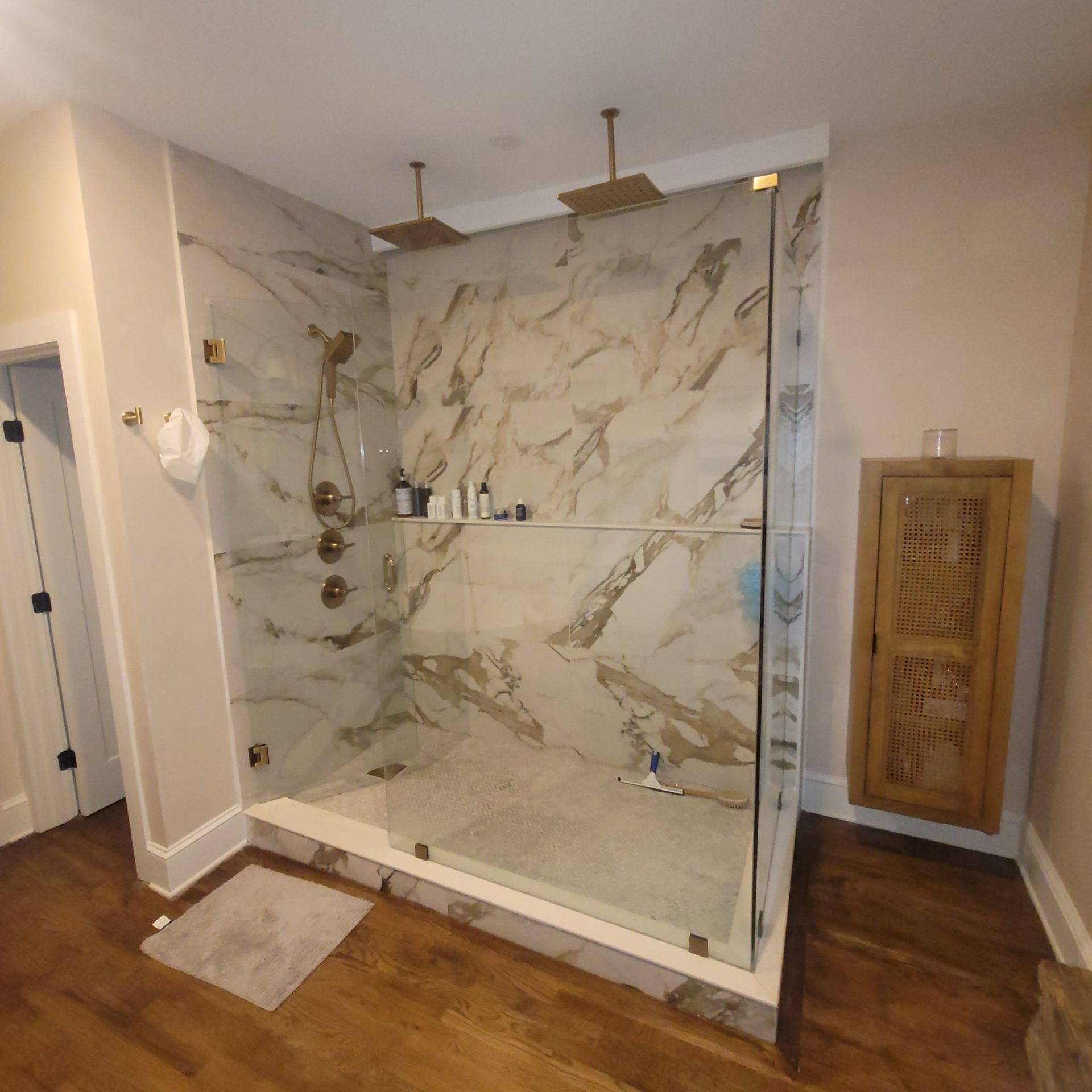 Luxurious marble-tiled shower with glass doors and two overhead showerheads, gold fixtures, and a wooden cabinet.