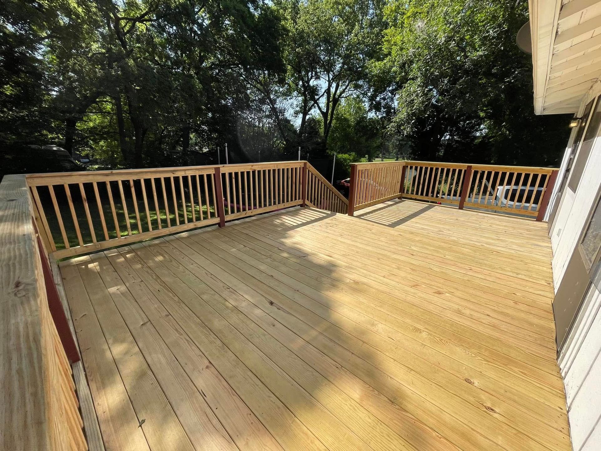 Wooden deck with railing, viewed from the corner of a house, surrounded by trees.