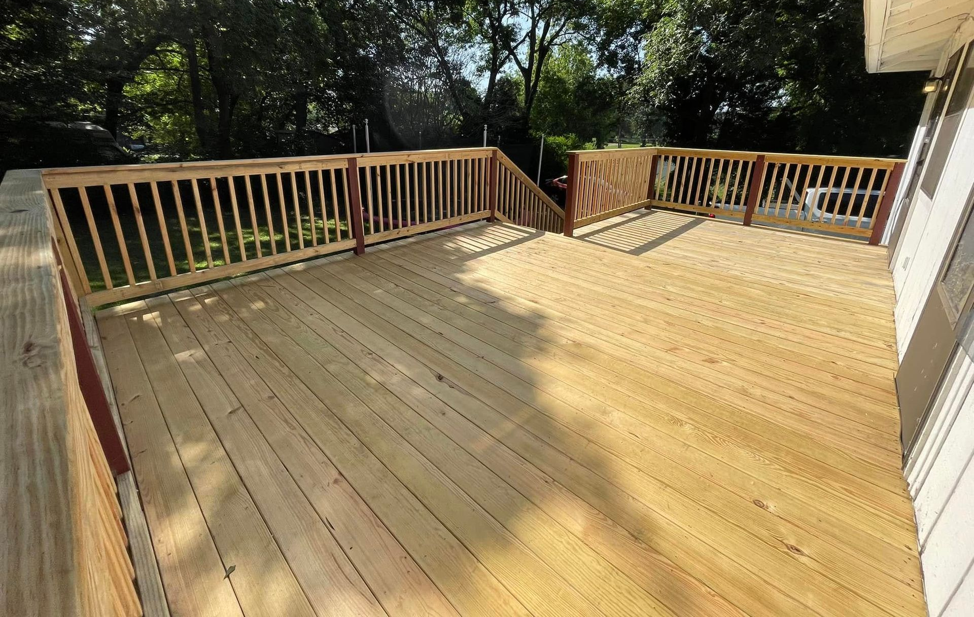 Newly built wooden deck with railing, surrounded by trees and attached to a house.