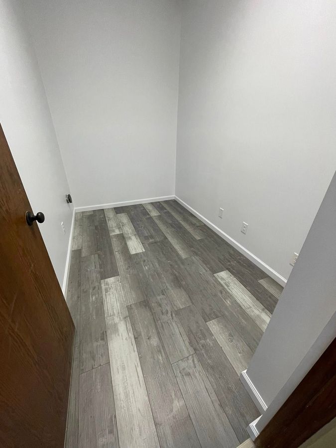 Small, empty room with gray wooden floor and white walls, brown door on the left.