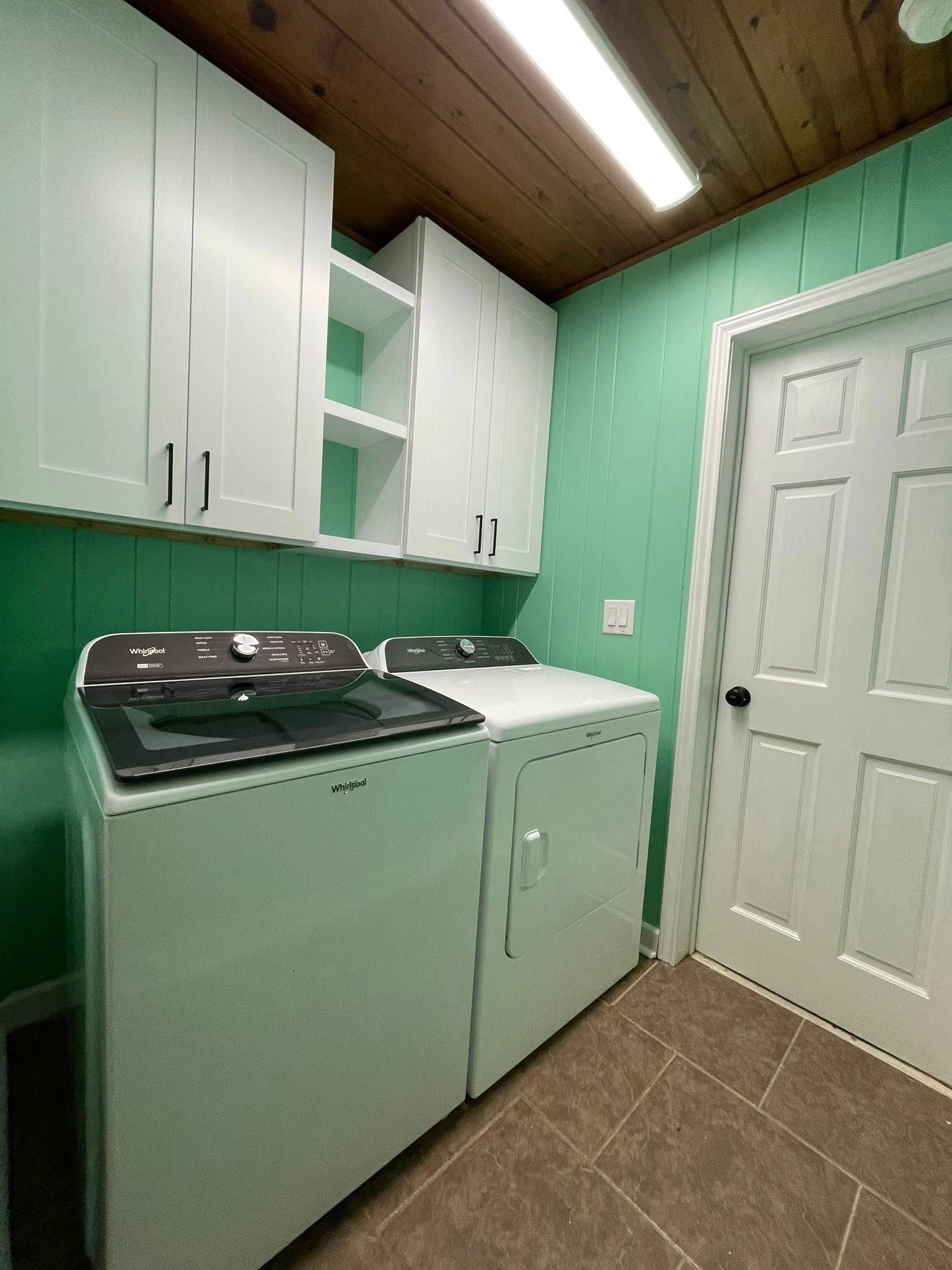 Laundry room with white washer and dryer, cabinets, and green walls. A closed white door is on the right.