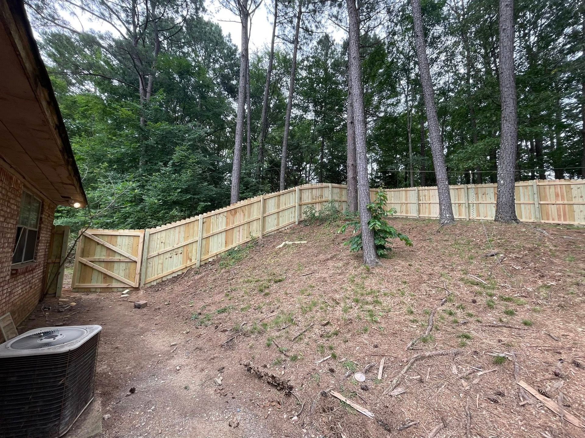 Wooden fence surrounds a backyard with a gate, trees, and a brick house.