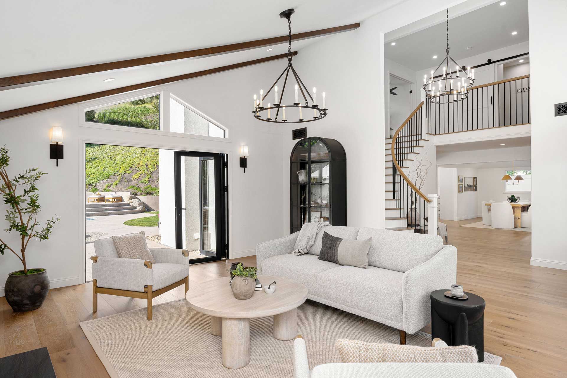 Living room with white walls, wooden beams, a sofa, armchairs, and a chandelier. A staircase leads to an upper level.