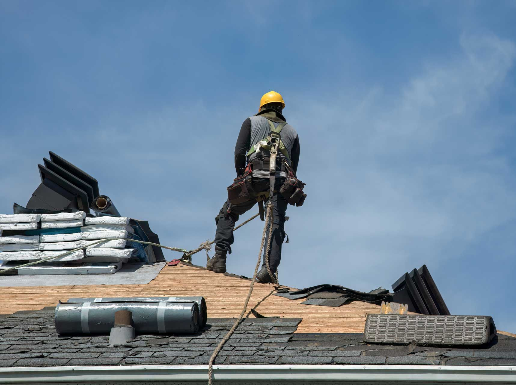 Roofer in safety harness on a partially stripped roof under a blue sky.