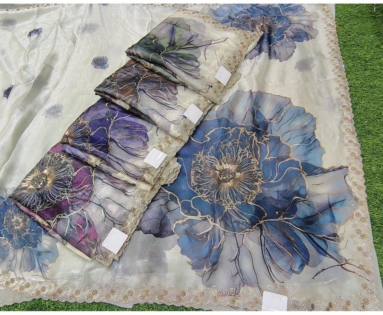 Folded fabric displays with large floral designs in blue, purple, and gold on a white background, arranged on grass.