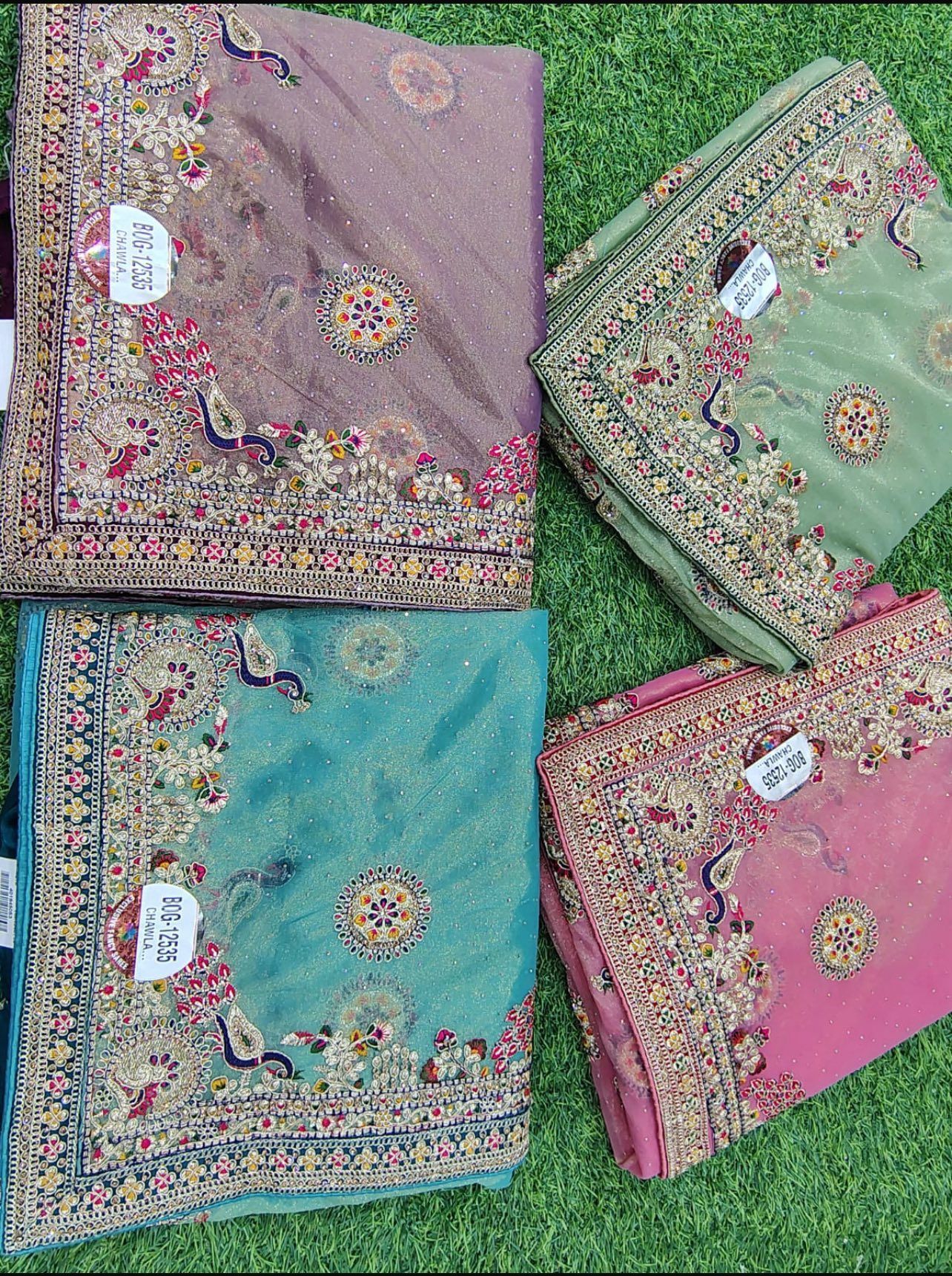 Four folded, intricately embroidered sarees in shades of purple, green, turquoise, and pink, on green grass.
