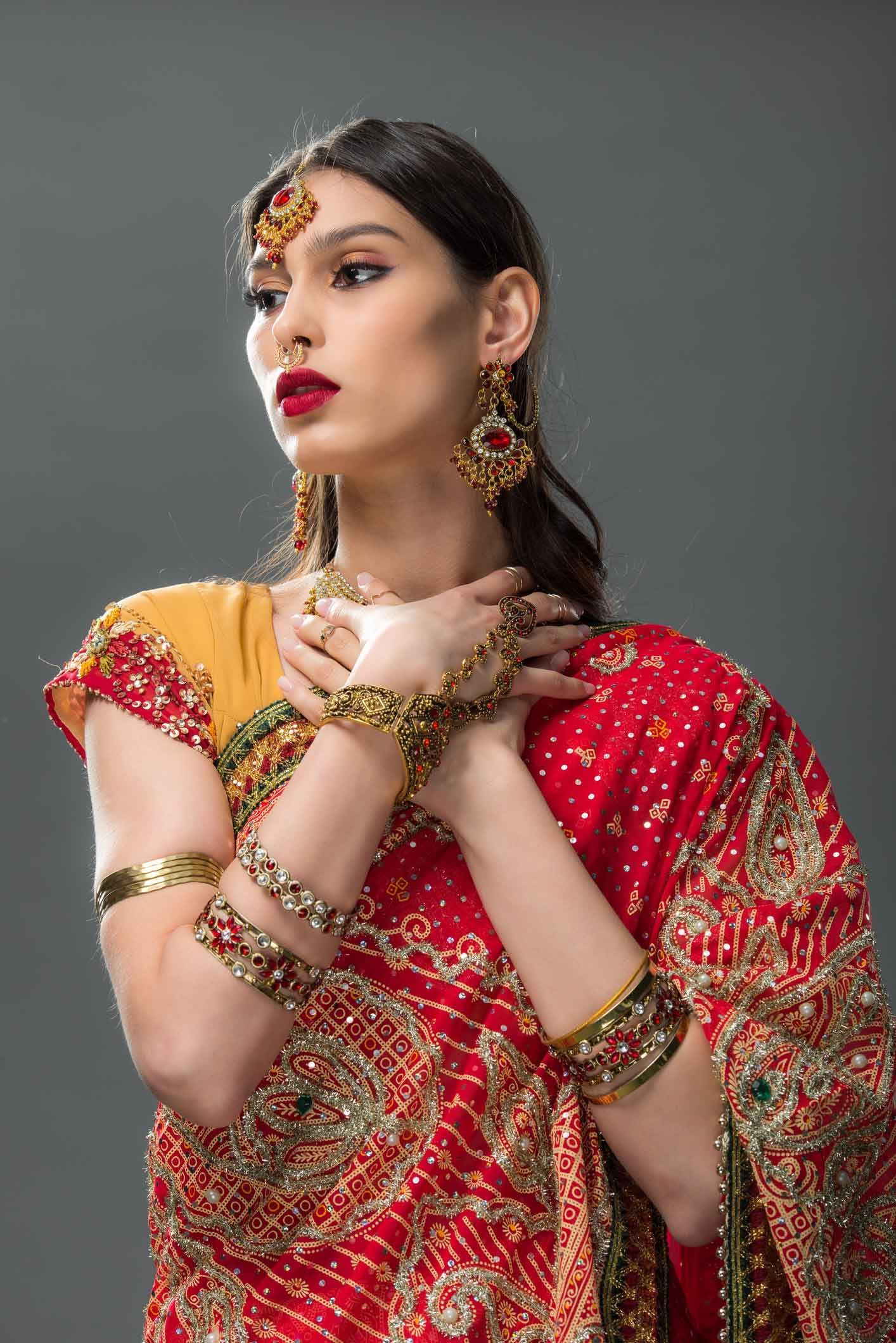 Woman in red sari, gold jewelry, and makeup, posing against a gray background.