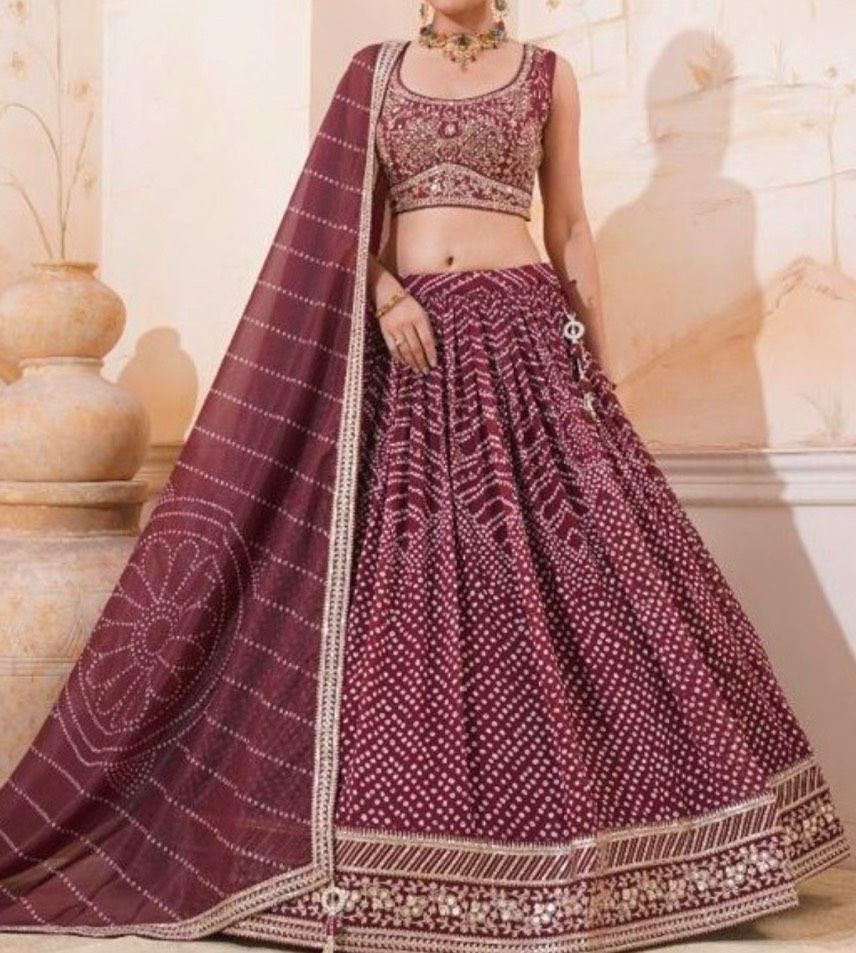 Woman wearing a maroon bandhani lehenga with a matching dupatta. She is in a beige-toned setting with ornate pottery.