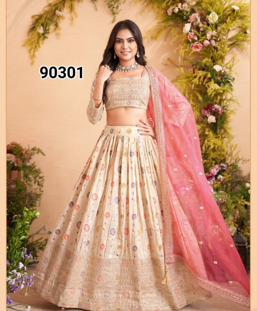 Woman in a cream and gold lehenga with a pink dupatta, posing in front of floral decorations. She is smiling and wearing a necklace.