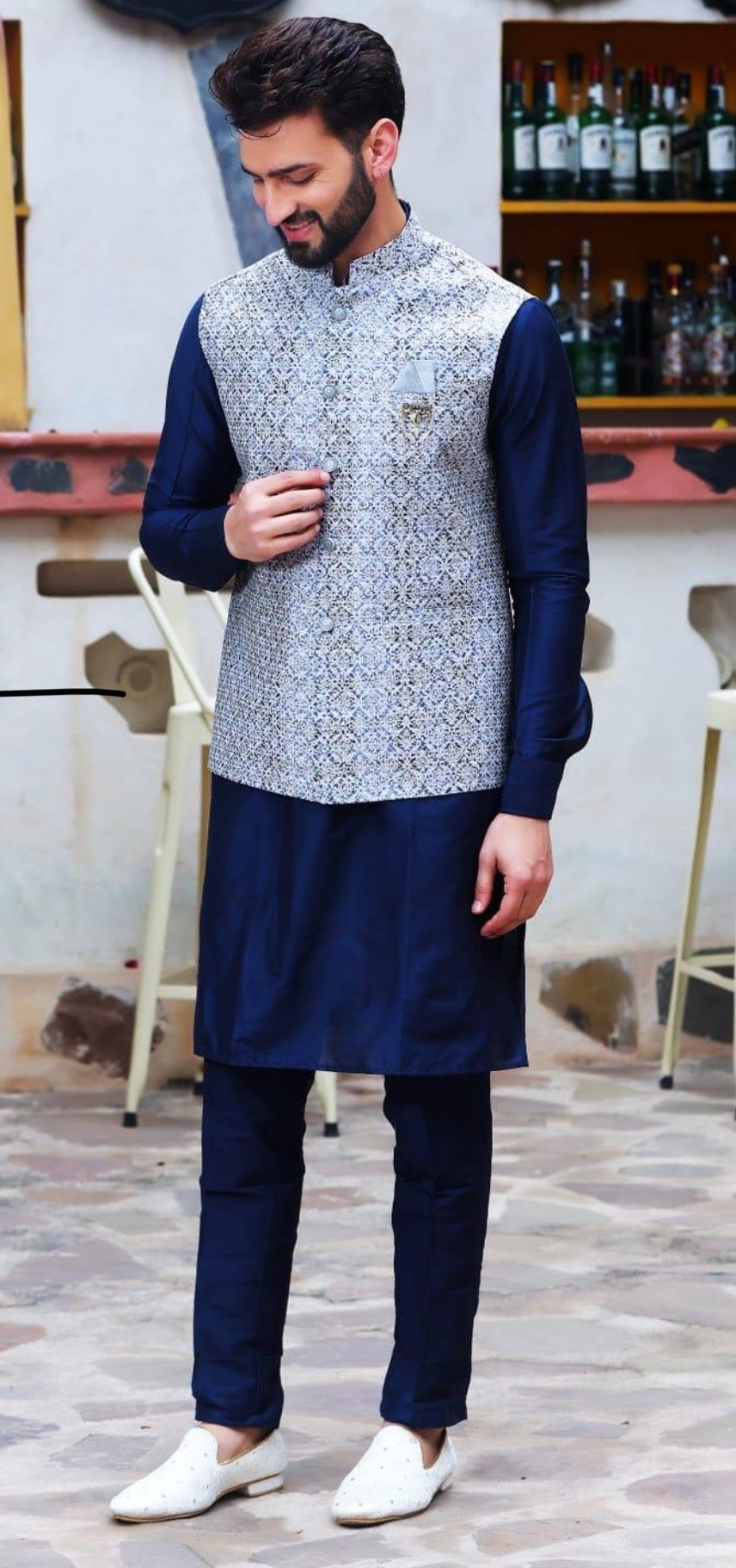 A man in a navy kurta, pants, and a patterned vest smiles. He's outside in a setting with bar stools and shelves of bottles.