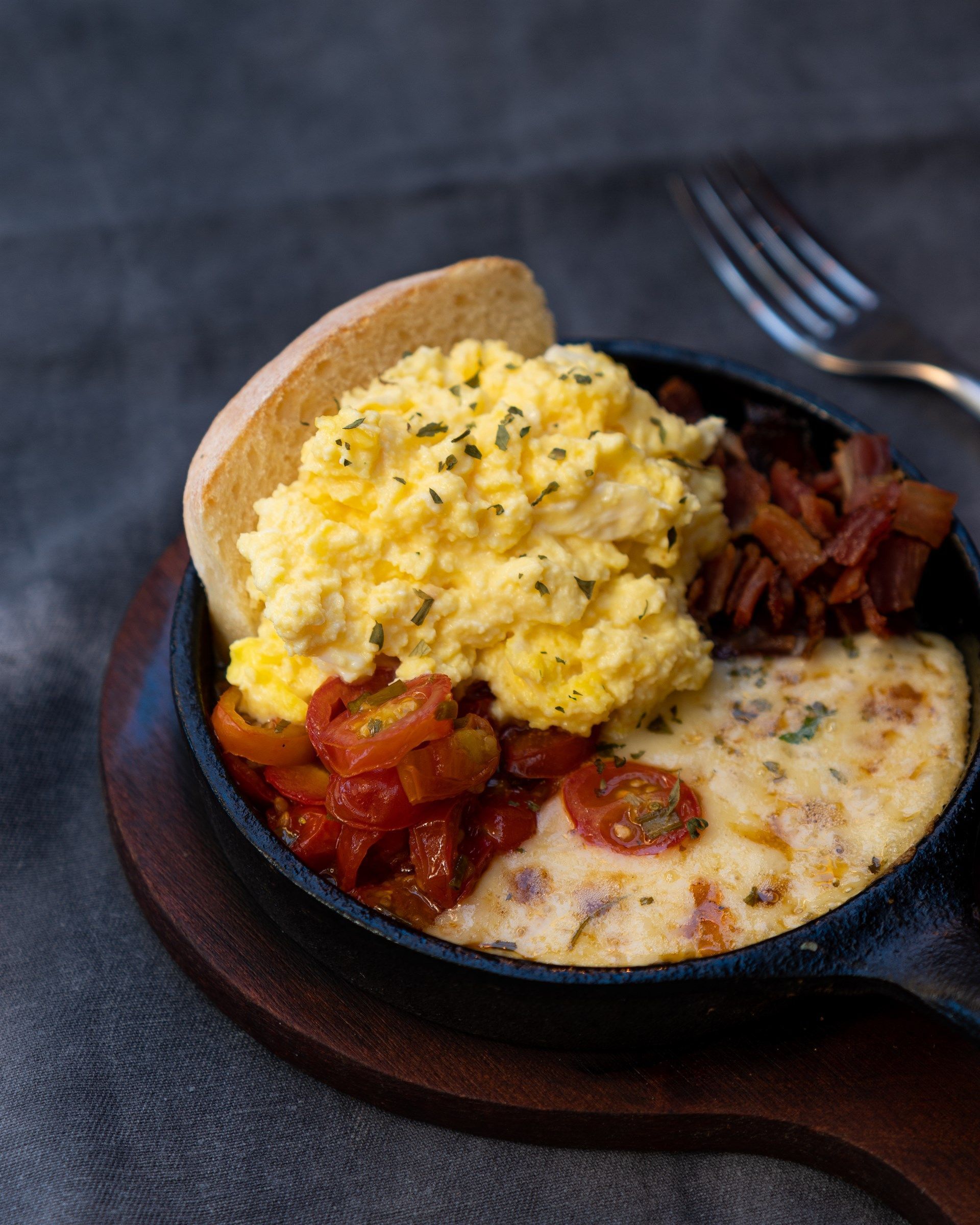 Una sartén llena de huevos revueltos, tomates, tocino y pan sobre una tabla de cortar de madera.
