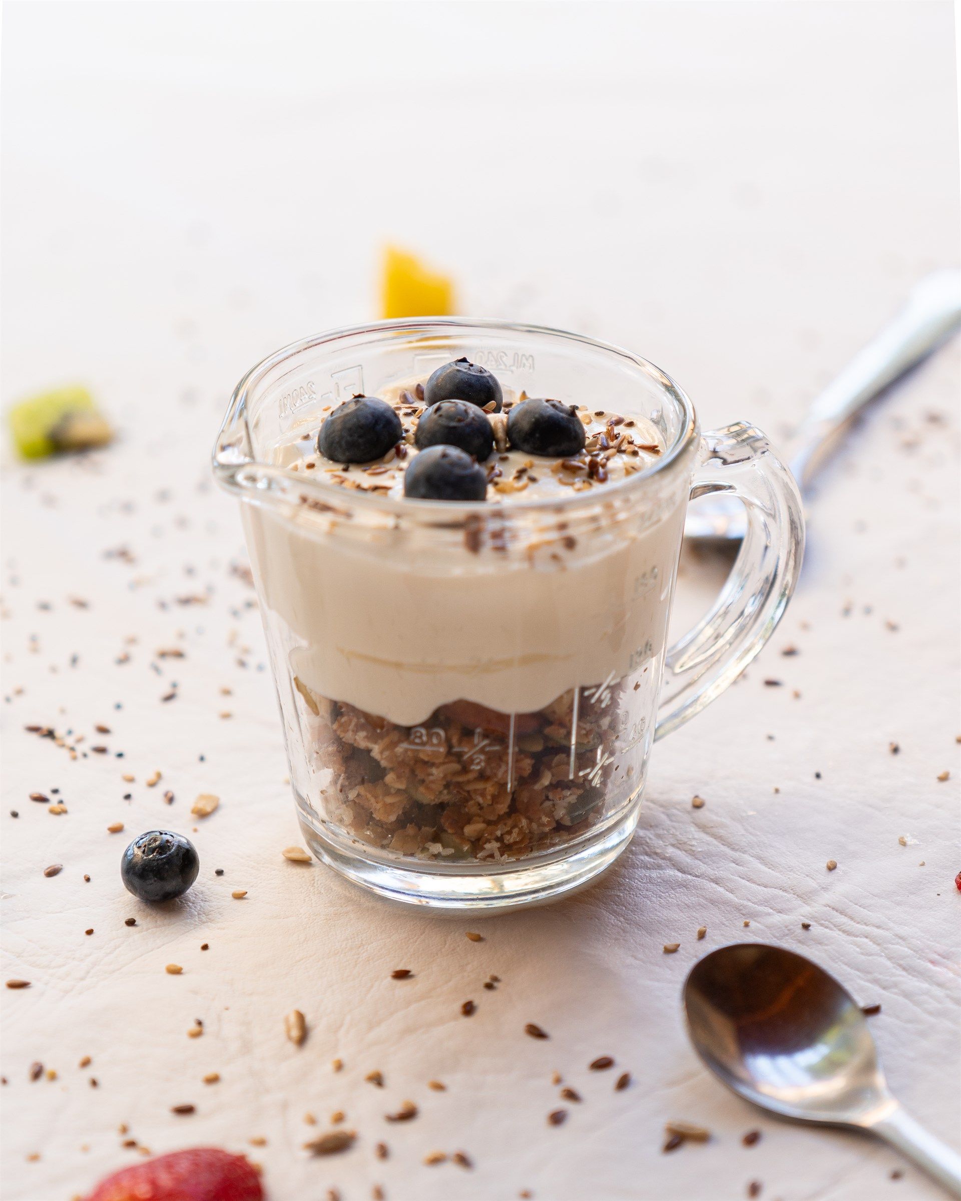 Un vaso de cristal lleno de yogur y granola al lado de una cuchara.
