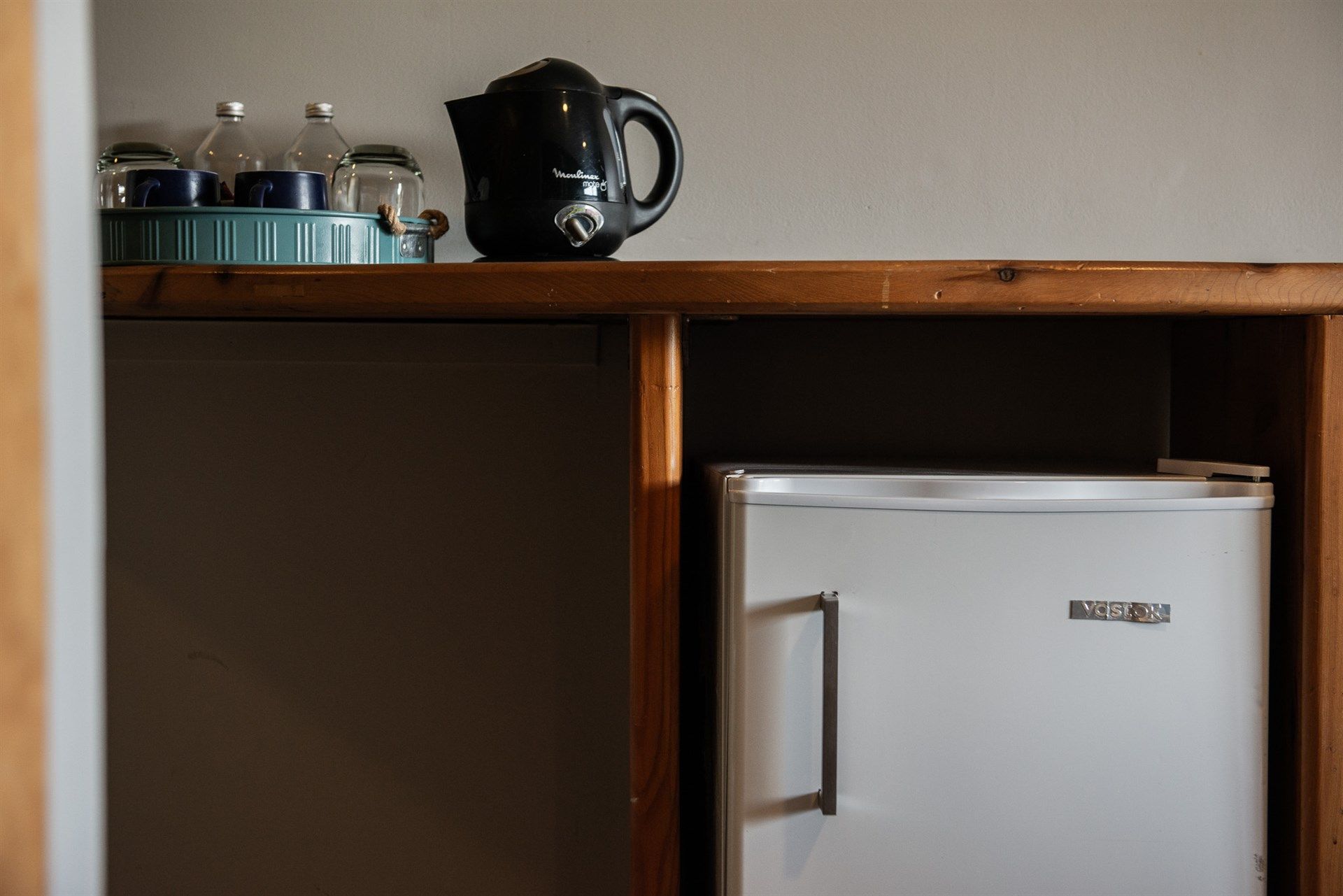 Debajo de una mesa de madera hay un pequeño frigorífico junto a una tetera.
