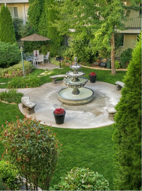 Exterior view of River Arms Apartments with three benches around water fountain and patio table with four chairs and umbrella.