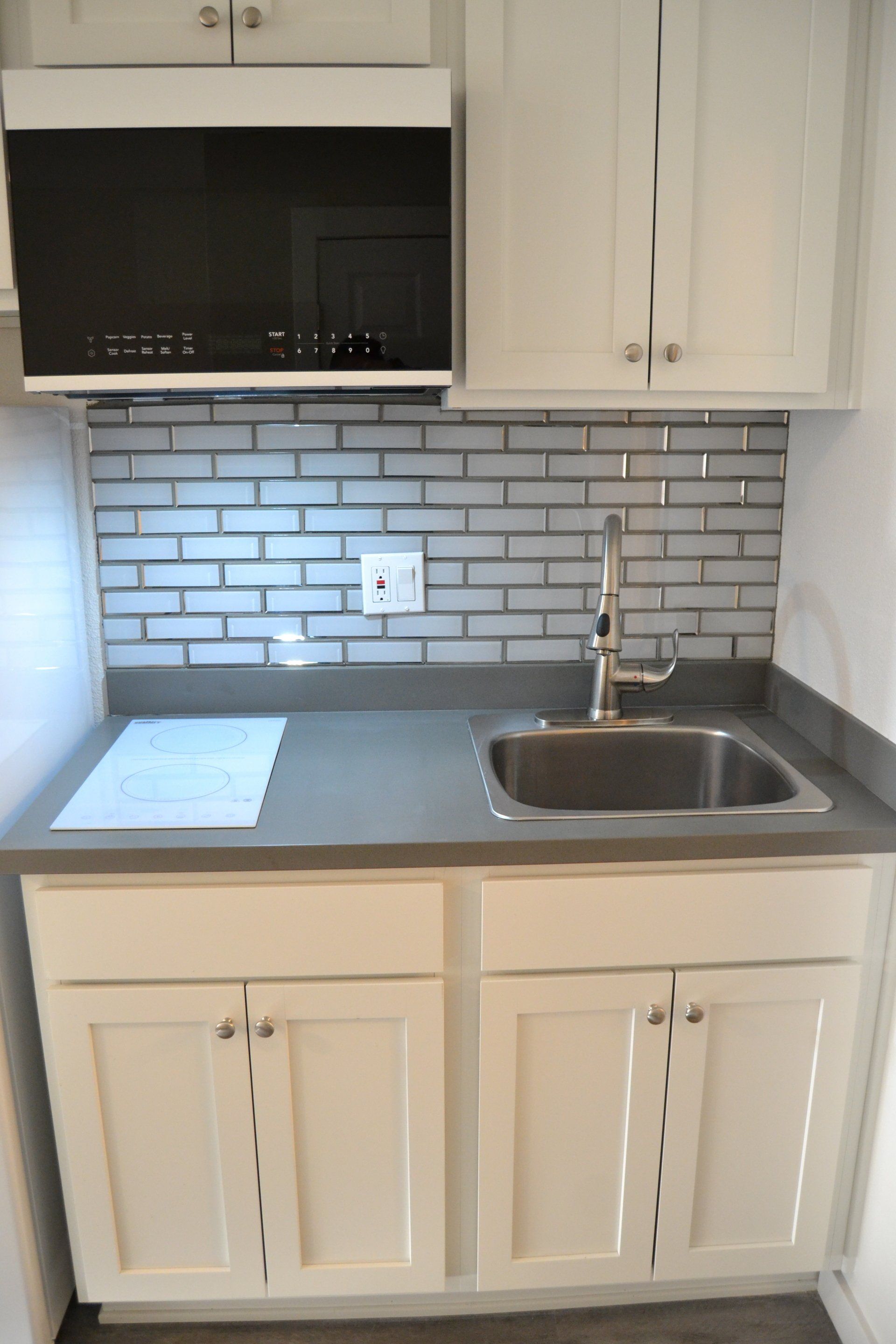 Updated studio kitchen with grey flooring, white cabinets, grey countertops, black microwave, white two electric burner stove, and white refrigerator.