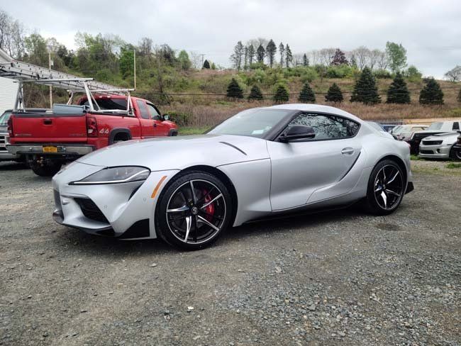 Silver Toyota Supra sports car parked outside. Red truck and other vehicles are in the background.