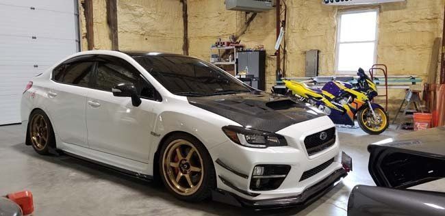 White Subaru sedan with bronze rims, carbon fiber hood, parked in a garage. A motorcycle sits behind it.