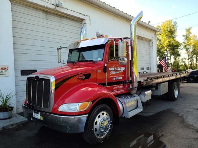 Red Peterbilt tow truck parked outside a building with garage doors, sunlight.