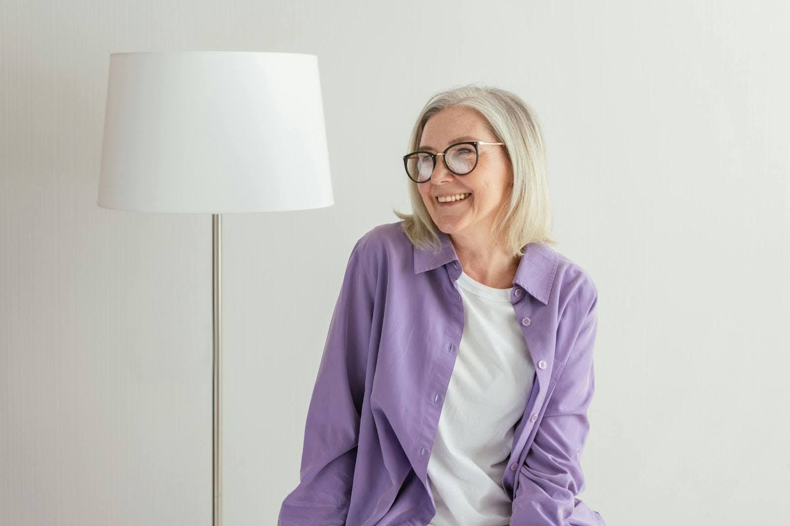 A woman wearing glasses and a purple shirt is sitting next to a lamp .