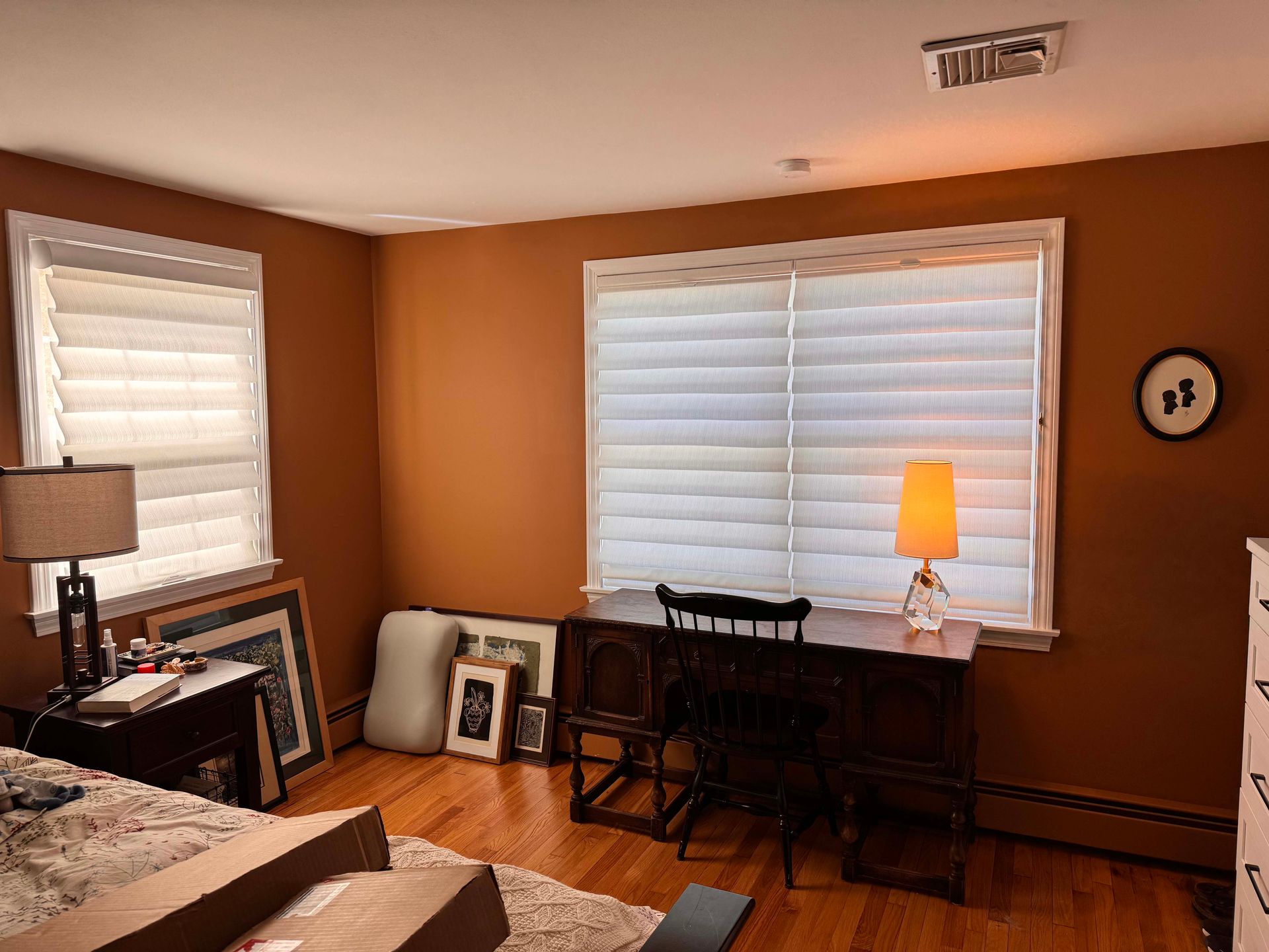 A living room with a desk , chairs , a lamp and two windows.