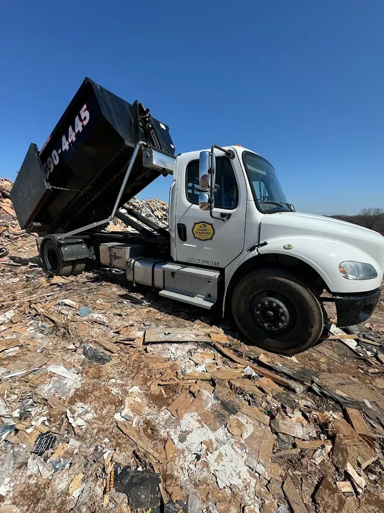 White dump truck on a rocky slope under a clear blue sky, truck bed raised and tilted.
