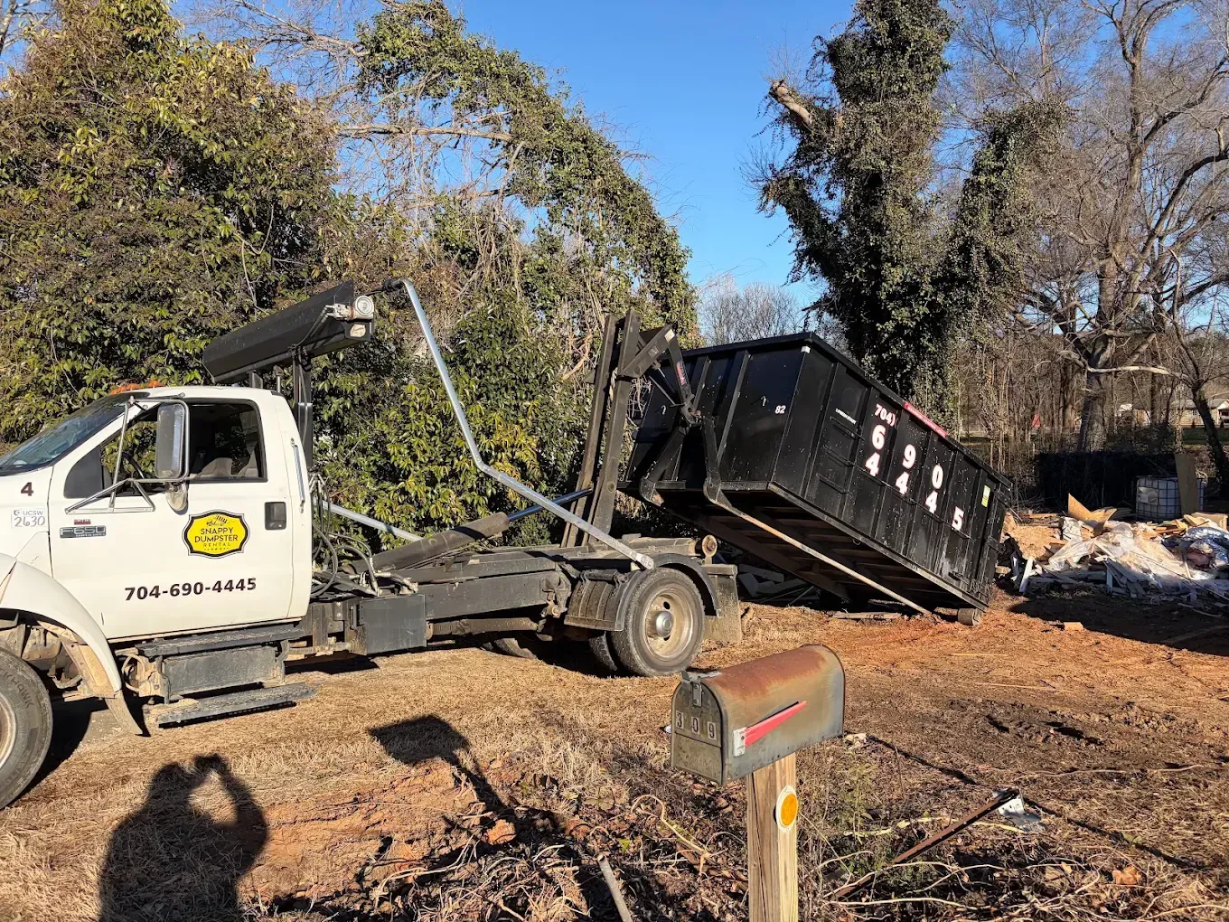 Tow truck lifting a black dumpster in a wooded area with debris and a mailbox nearby