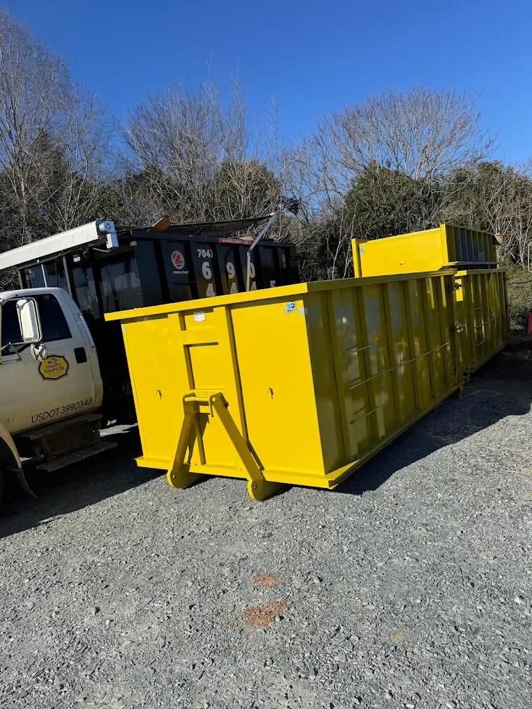 Yellow construction dumpster beside a utility truck on a gravel lot under a clear blue sky