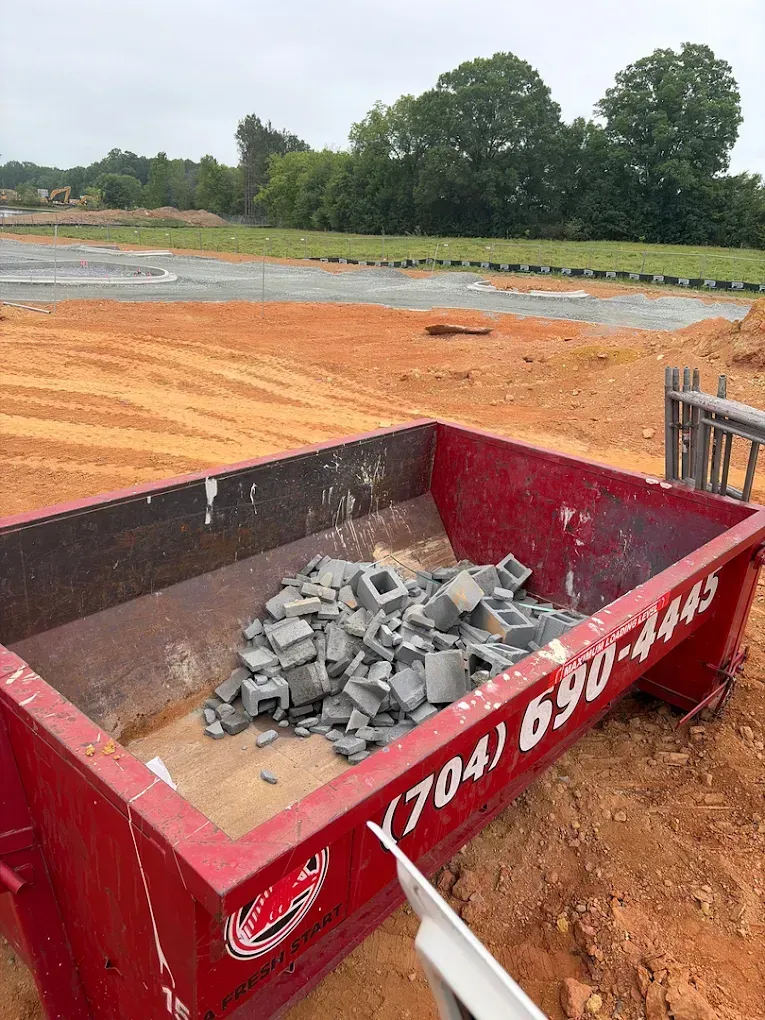 Red construction dumpster filled with gray rubble on an orange dirt site near trees and open ground
