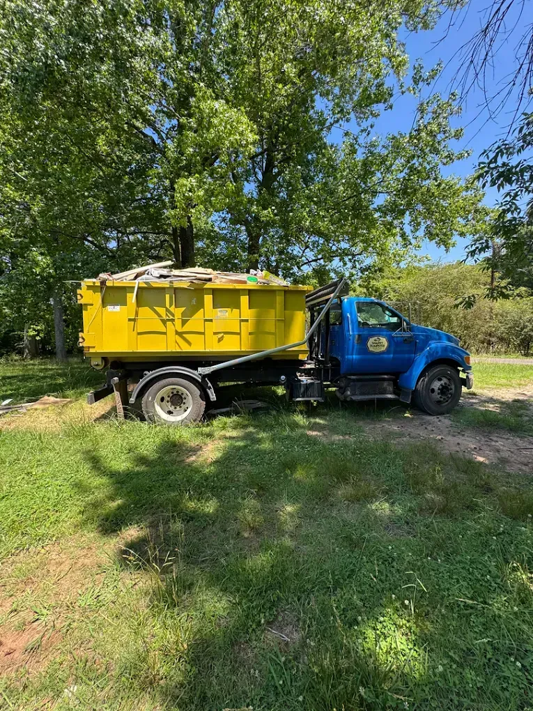 Blue dump truck with a yellow bed parked on grass under leafy trees in sunlight