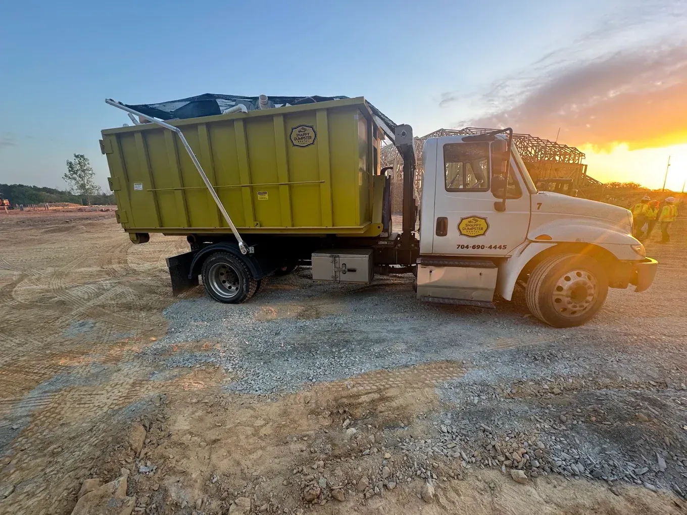 Yellow dump truck at sunset on a gravel lot, with a large green cargo bed.