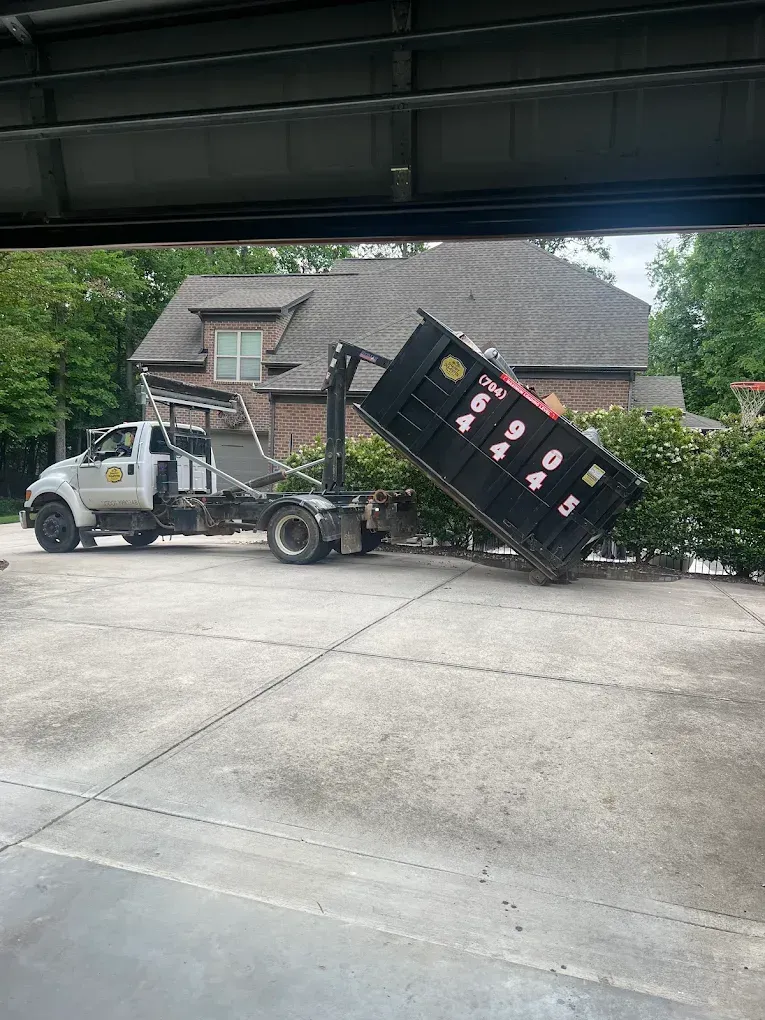 Dump truck tipping a black dumpster in a suburban driveway, with a house and trees in the background.