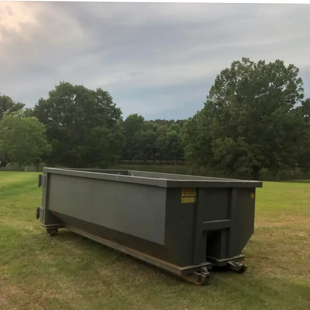 Large dark gray industrial dumpster on grass in a field with trees and cloudy sky
