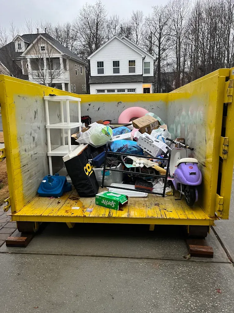 Yellow dumpster filled with mixed household trash on a street, with houses in the background
