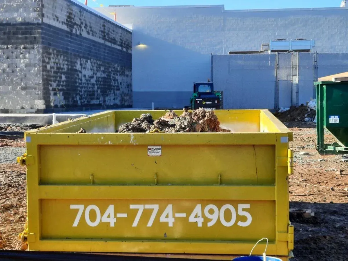 Yellow dumpster with debris in an industrial lot, marked 704-774-4905