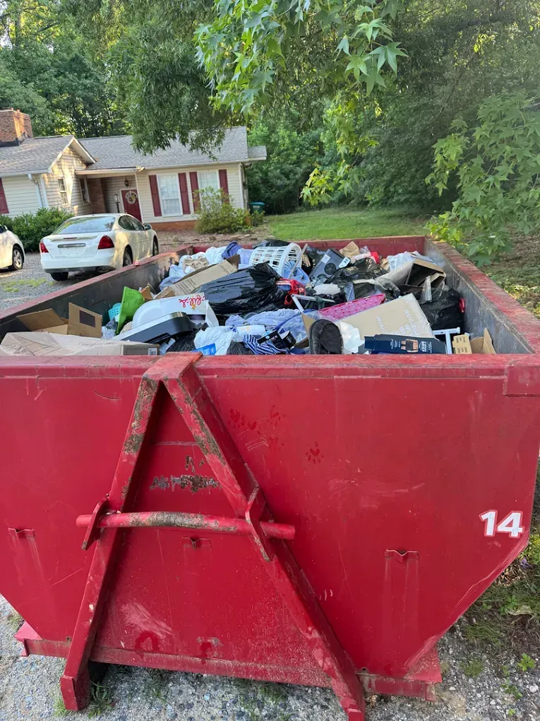 Red dumpster overflowing with assorted trash in a suburban driveway, with a white car and houses in the background