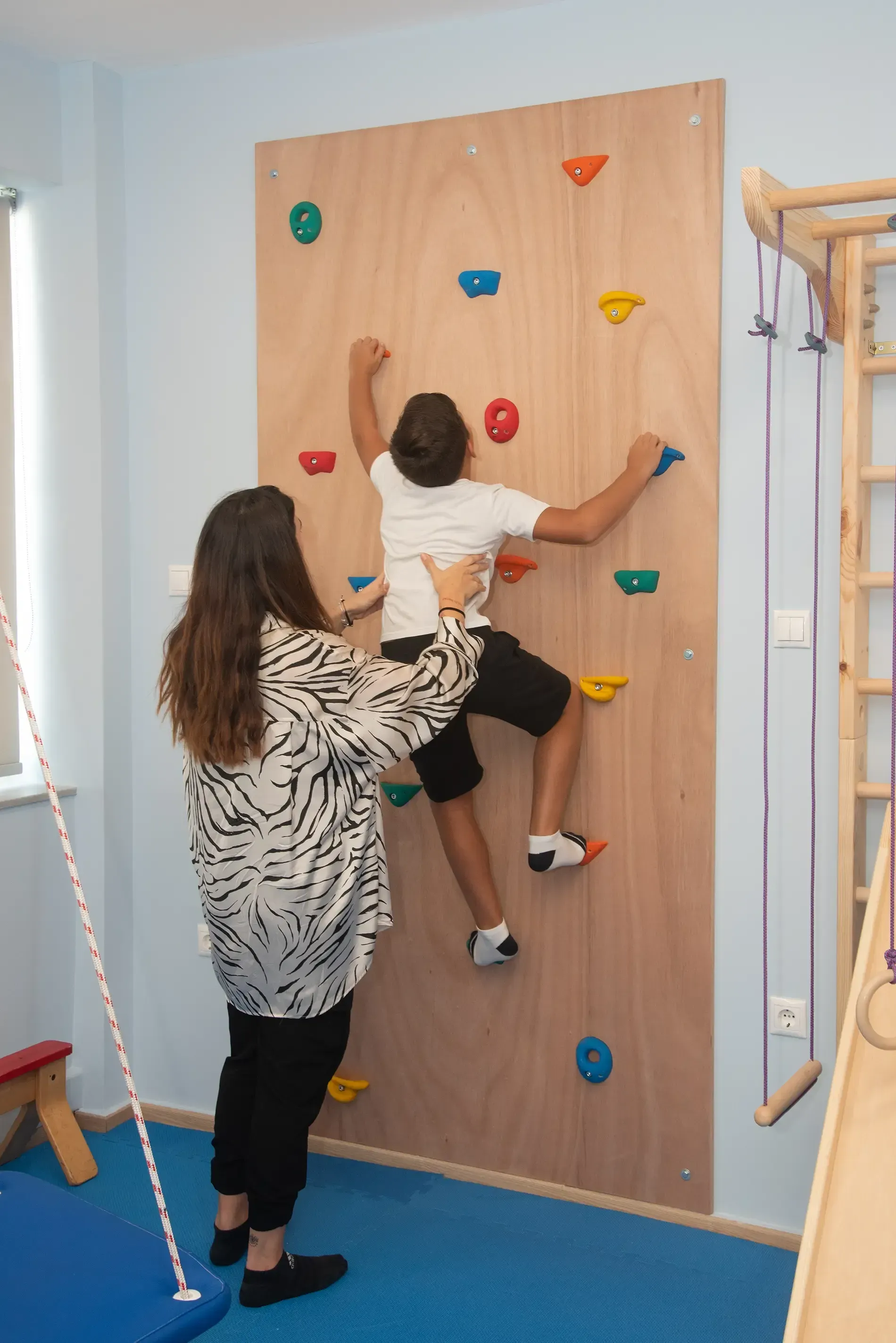 Μια γυναίκα βοηθά ένα νεαρό αγόρι να σκαρφαλώσει σε έναν τοίχο αναρρίχησης.