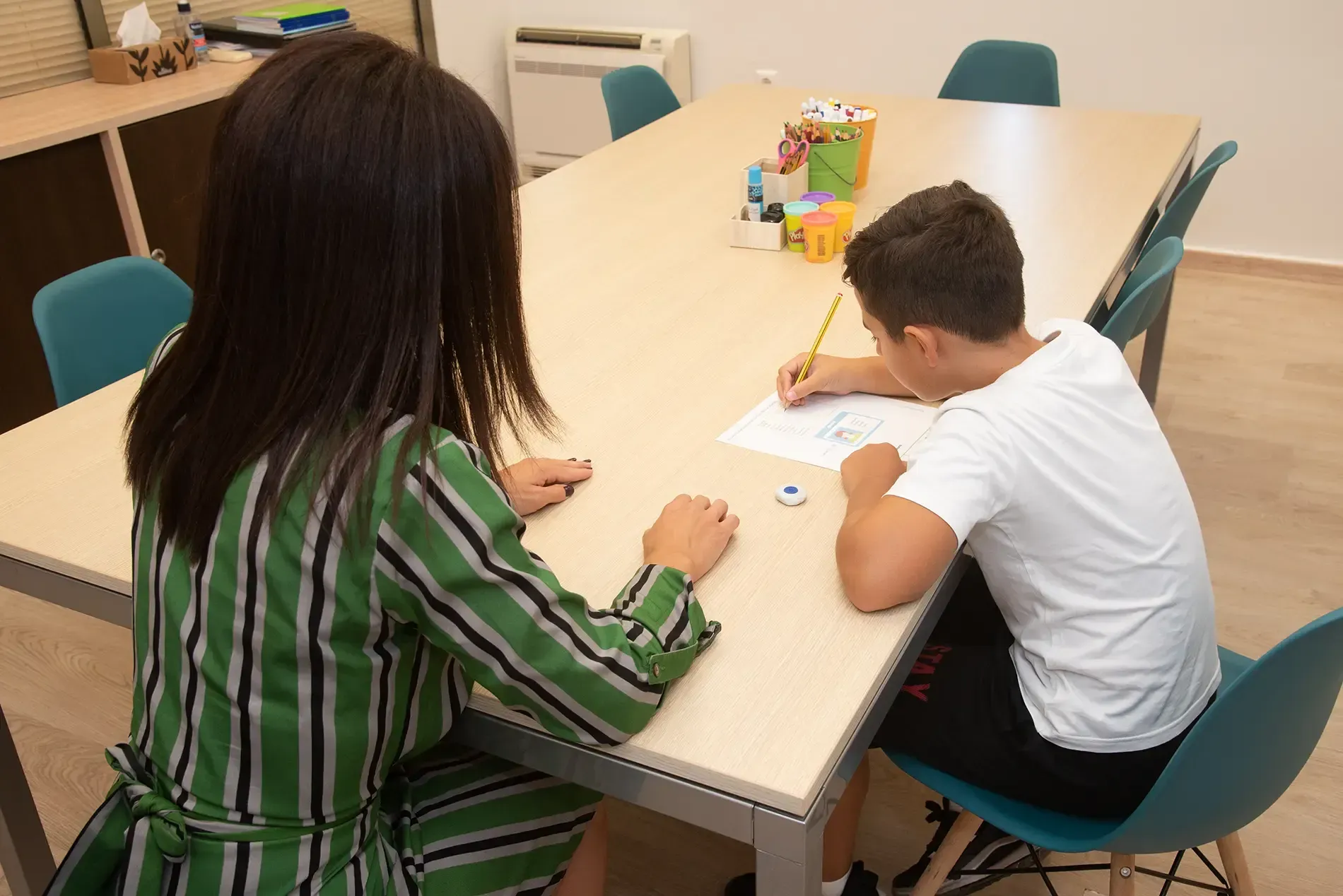 Μια γυναίκα κάθεται σε ένα τραπέζι και μιλάει σε ένα αγόρι που γράφει σε ένα κομμάτι χαρτί.