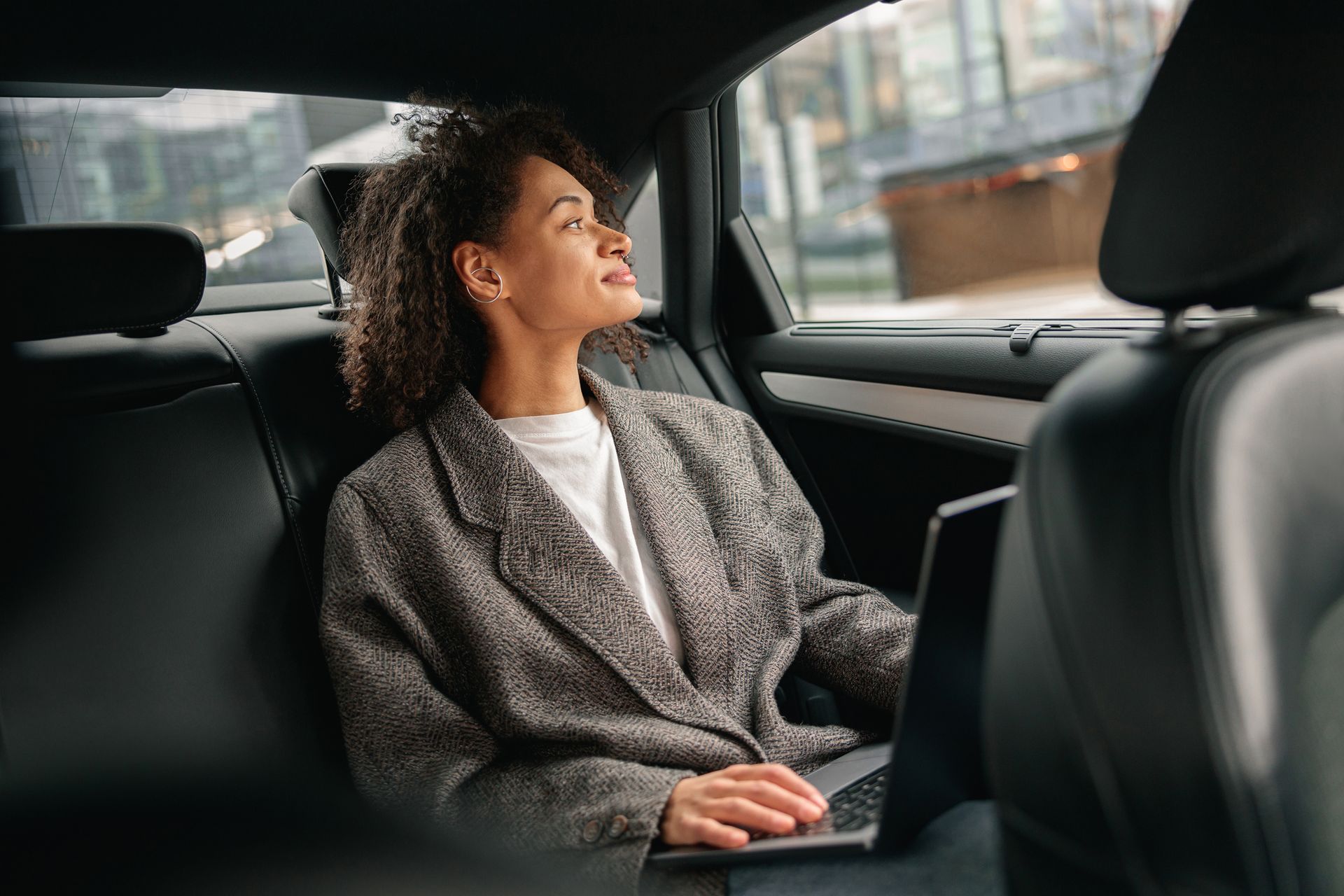 Woman in blazer looking out car window, laptop on lap, city backdrop.