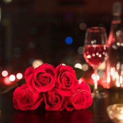 Red roses, a glass of wine, and a bottle sit on a dark surface; warm lighting in the background creates a romantic ambiance.