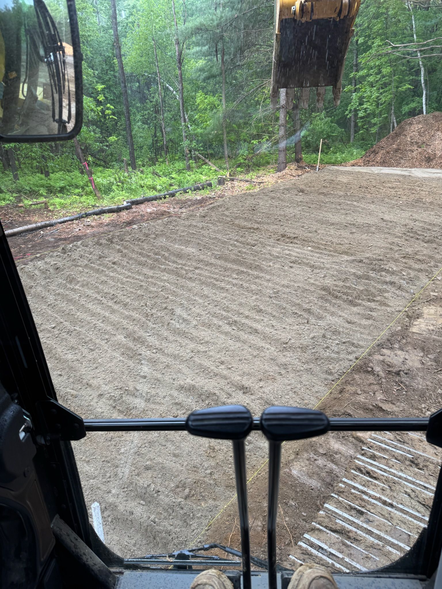 Inside an excavator cab, a freshly graded dirt path in a rainy wooded area with another machine arm visible.