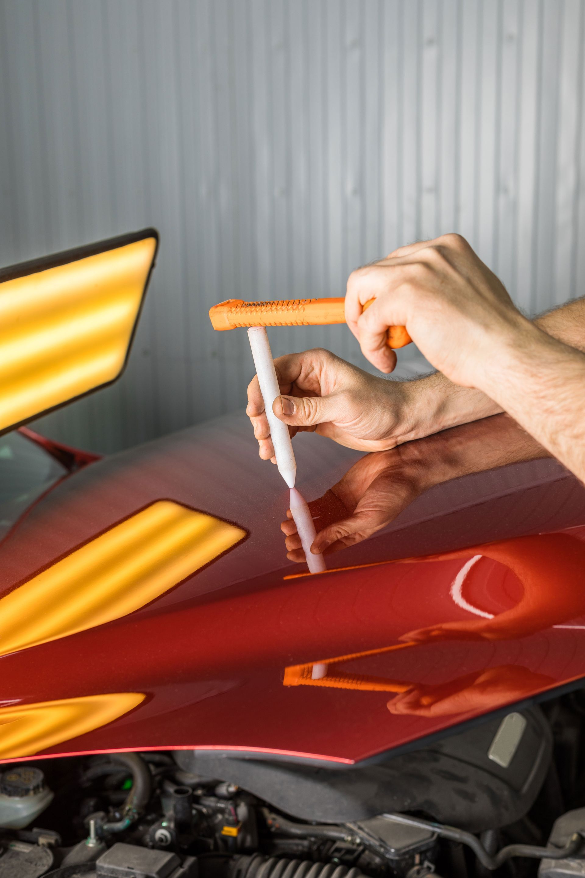 Person uses an orange handle tool to dent-pull a red car hood inside a garage.