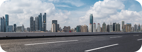 Uma estrada de asfalto com linhas brancas leva ao horizonte de uma cidade sob um céu azul com nuvens.