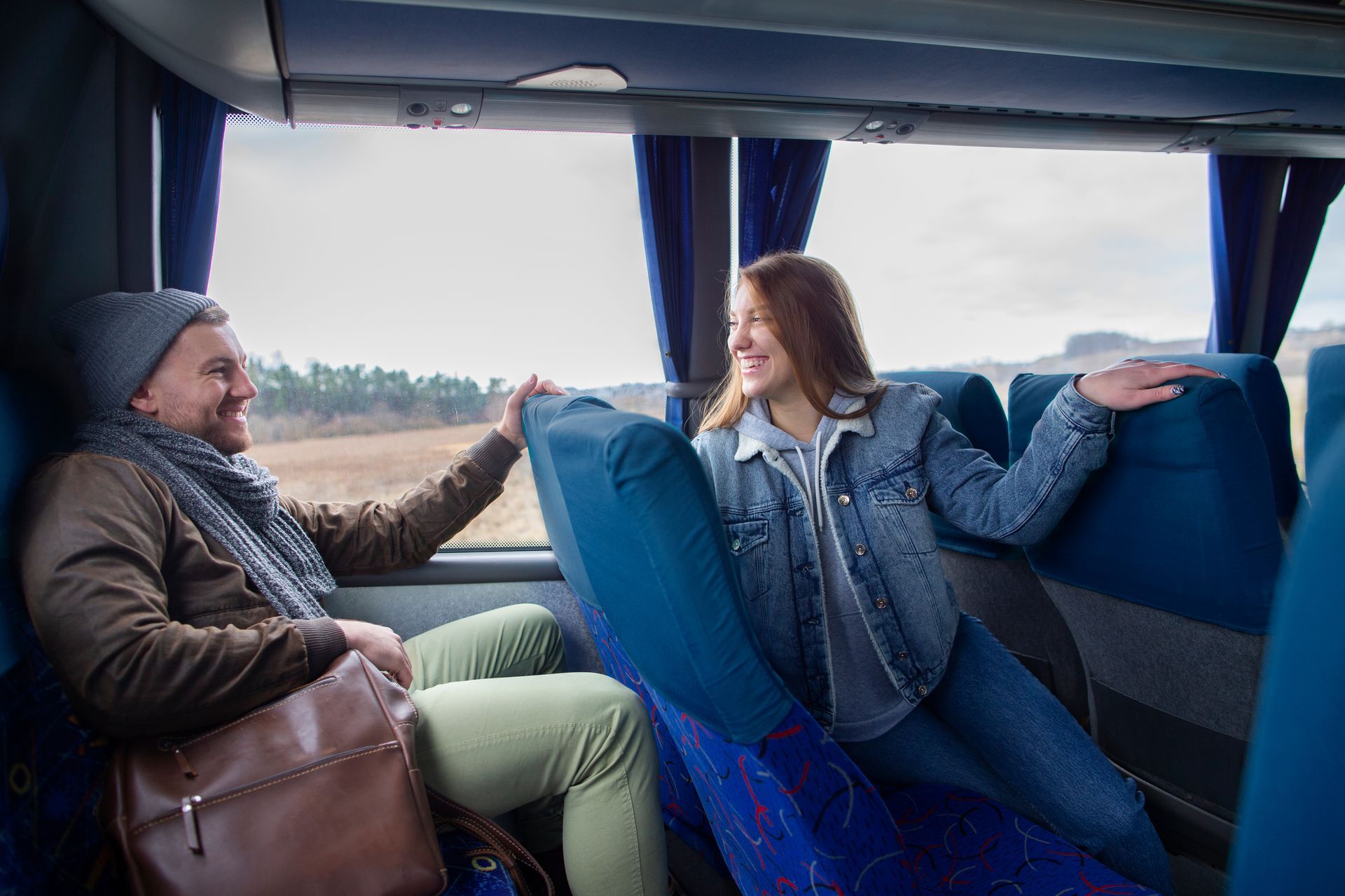 Homem e mulher sorrindo, estendendo as mãos um para o outro em um ônibus, próximos a janelas com vista para o exterior.