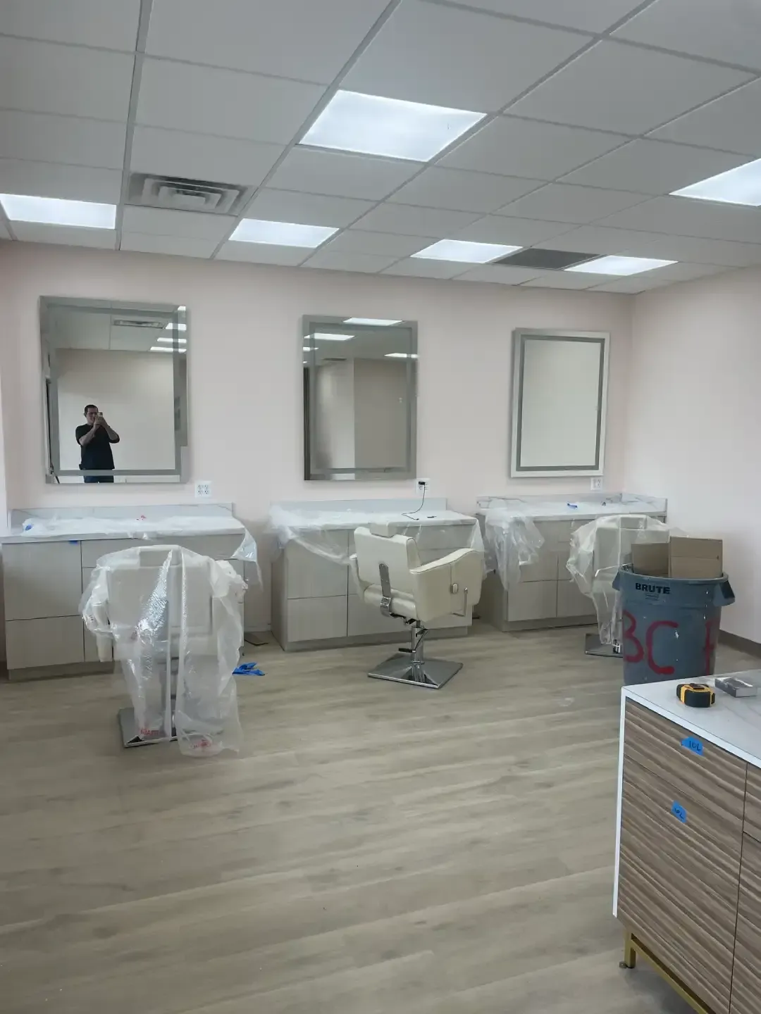 Empty salon room with mirrors, chairs wrapped in plastic, and a wood floor under fluorescent lights