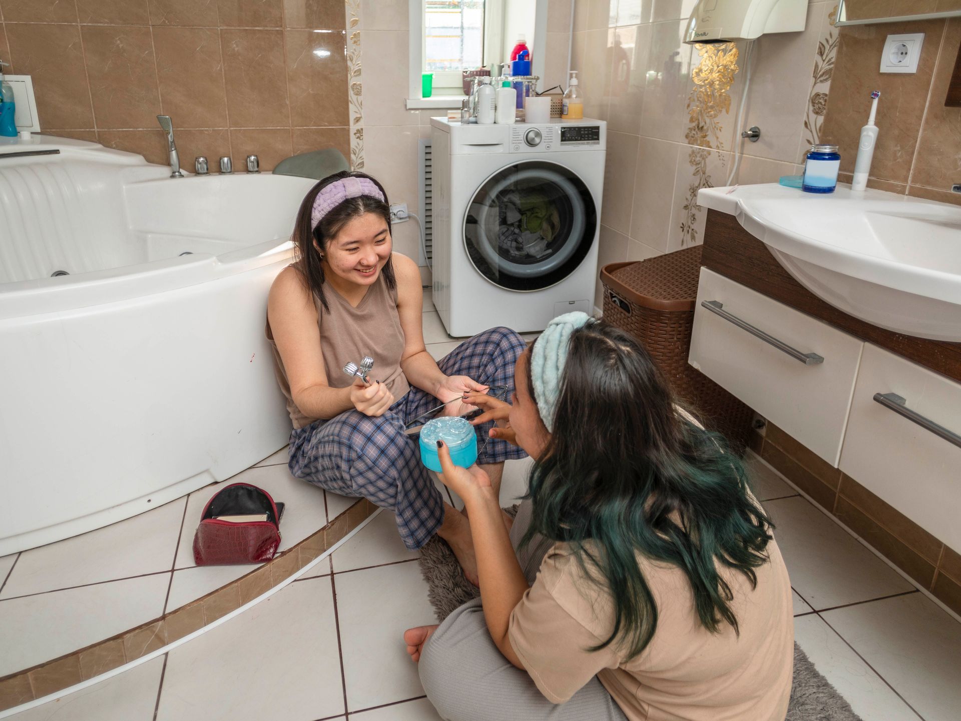 Two women are sitting on the floor in a bathroom next to a bathtub.