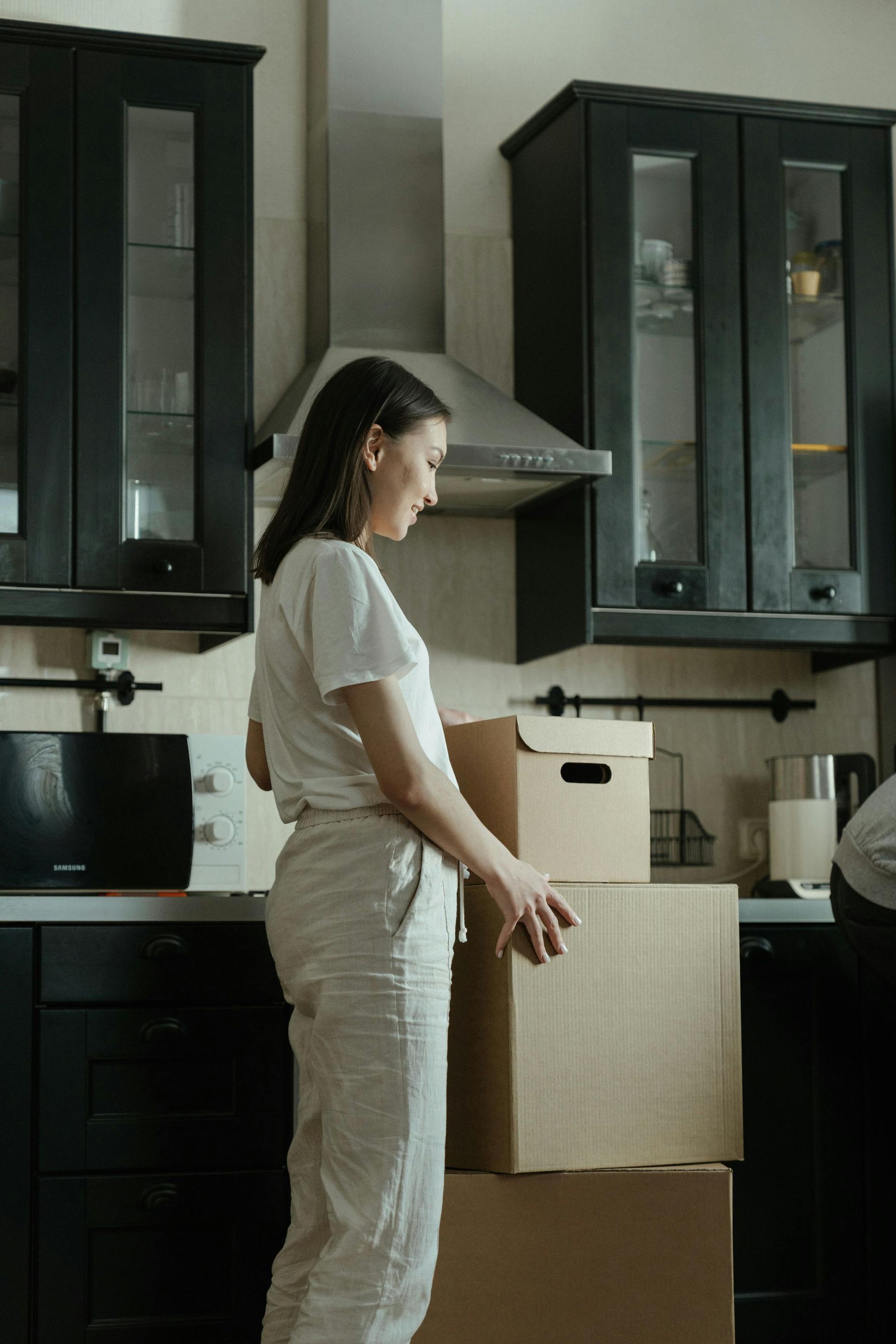 Woman in kitchen, moving boxes near cabinets and appliances.