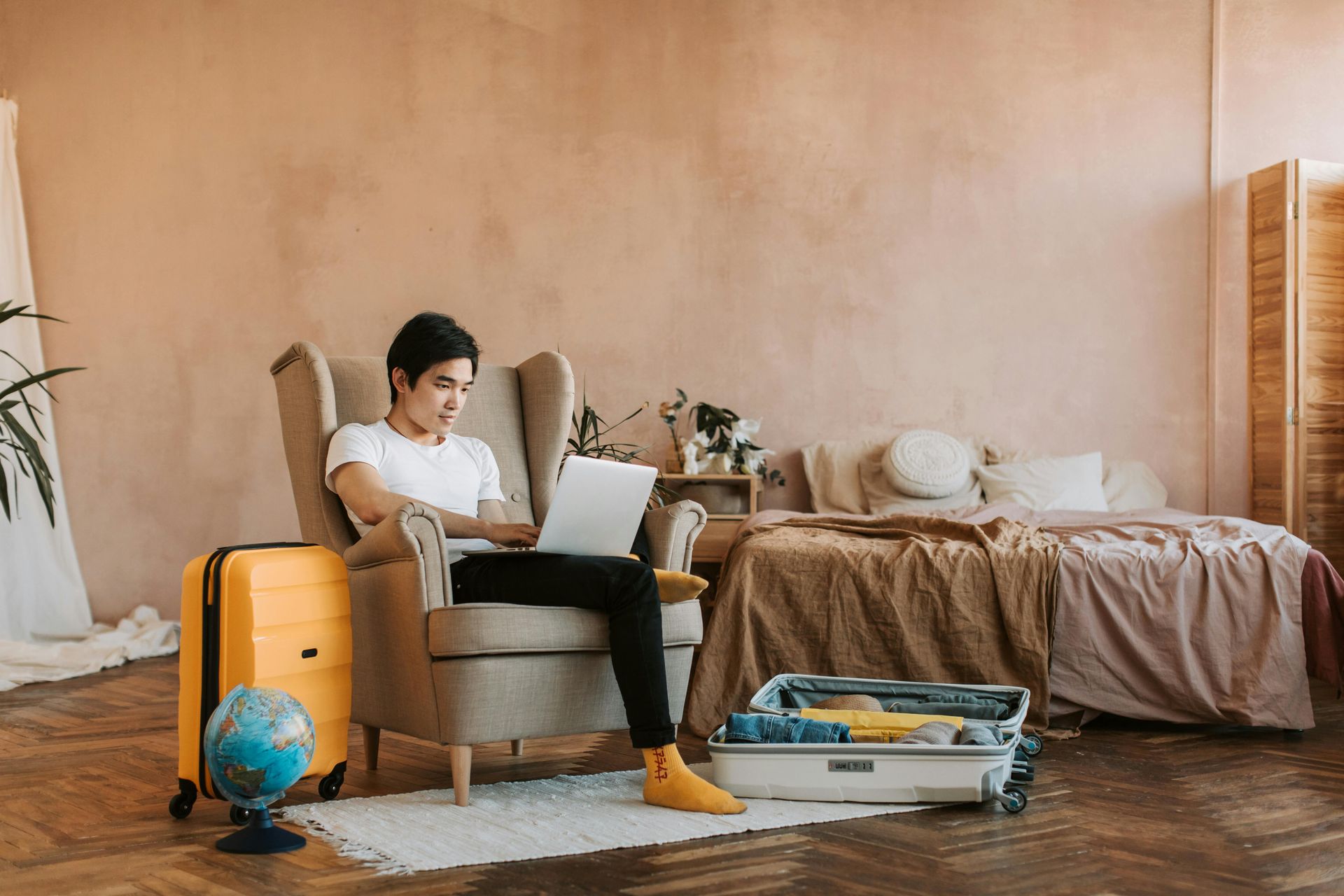 Man on laptop in a chair, luggage and bed nearby, globe on floor. Brown walls, natural light.