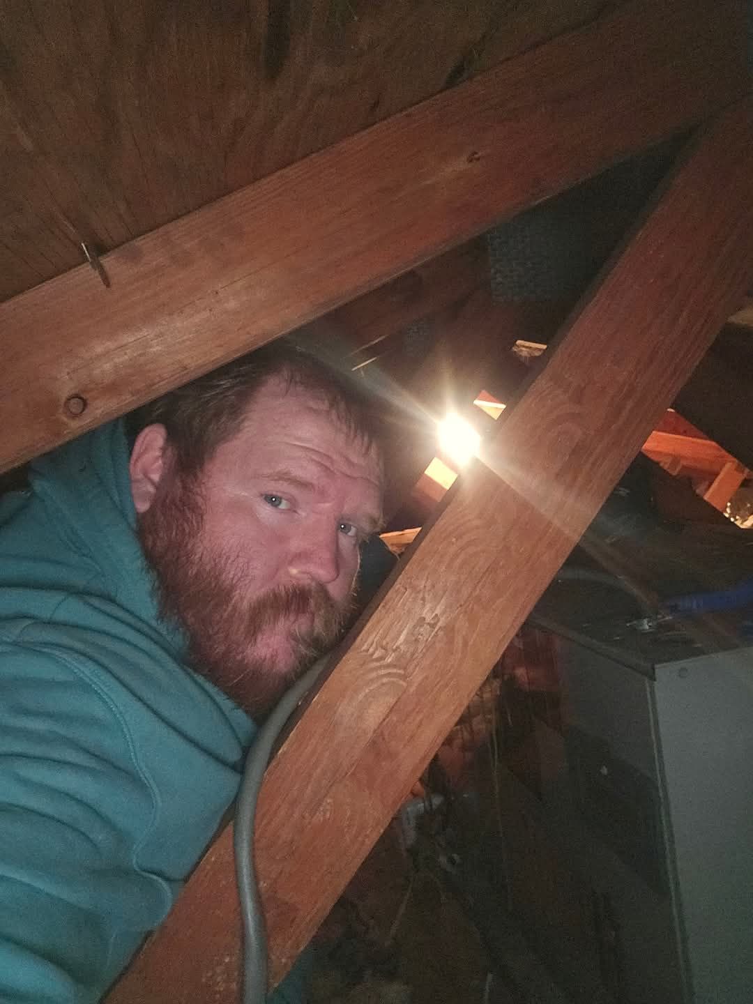 Man peering out from an attic space, lit by a bright light. He has a red beard and wears a blue hoodie.